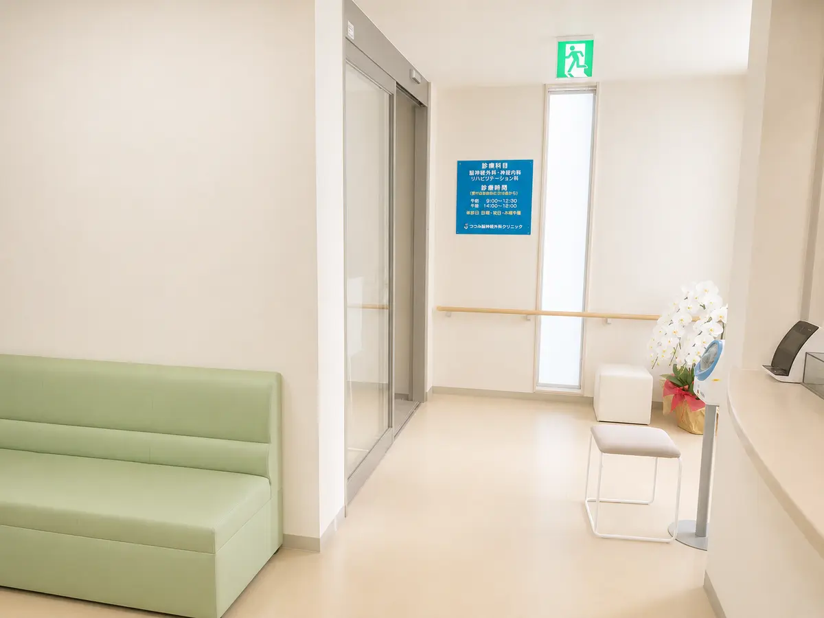 つつみ脳神経外科クリニックの入口。自動ドアの脇に淡い緑色のソファと、花を生けた鉢が置かれている。奥の壁には診療科目・診療時間を記した案内板がある。